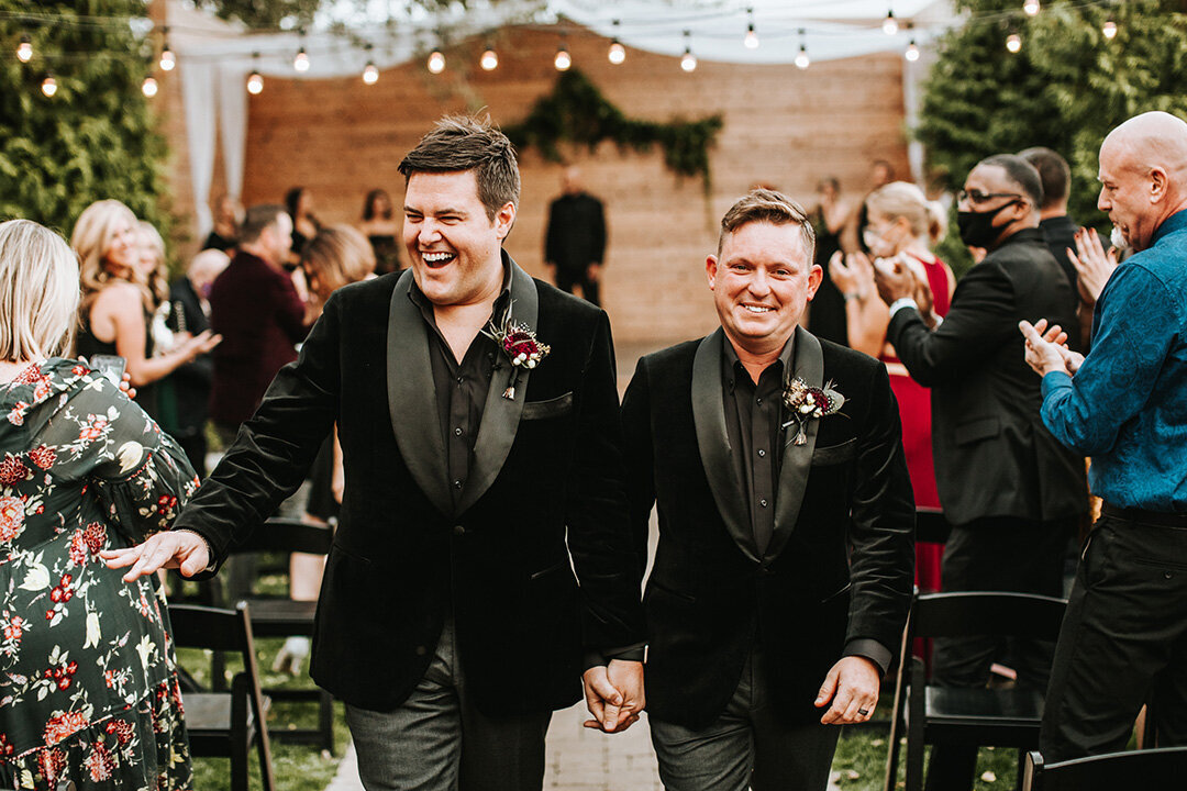 Two grooms wearing black tuxedos walk down the aisle holding hands.