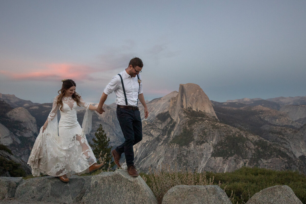 A groom walks in front of his bride holding her hand behind him along a rock walk in Yosemite National Park as the sunsets behind them.