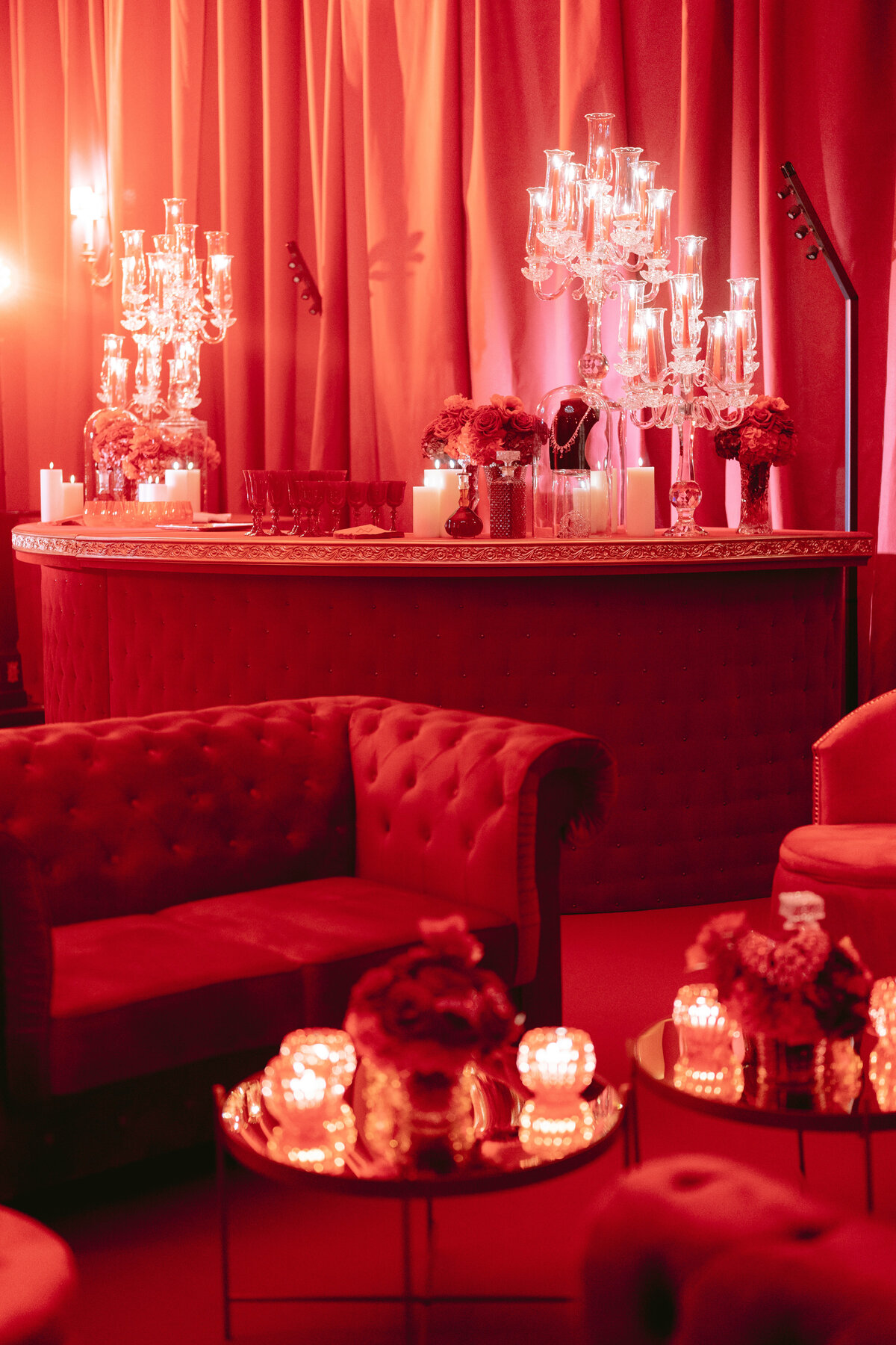 The photo you're referring to was taken at the Shangri La Hotel & Ritz Paris Hotel in Paris, France, capturing the exquisite work of photographer Matteo Coltro. An elegant lounge area bathed in red light is showcased, with tufted couches, ornate candles, and floral arrangements adorning the space. The bar is adorned with tall candelabras, and in the background, luxurious curtains complete the scene, adding to the intimate ambiance of the setting.