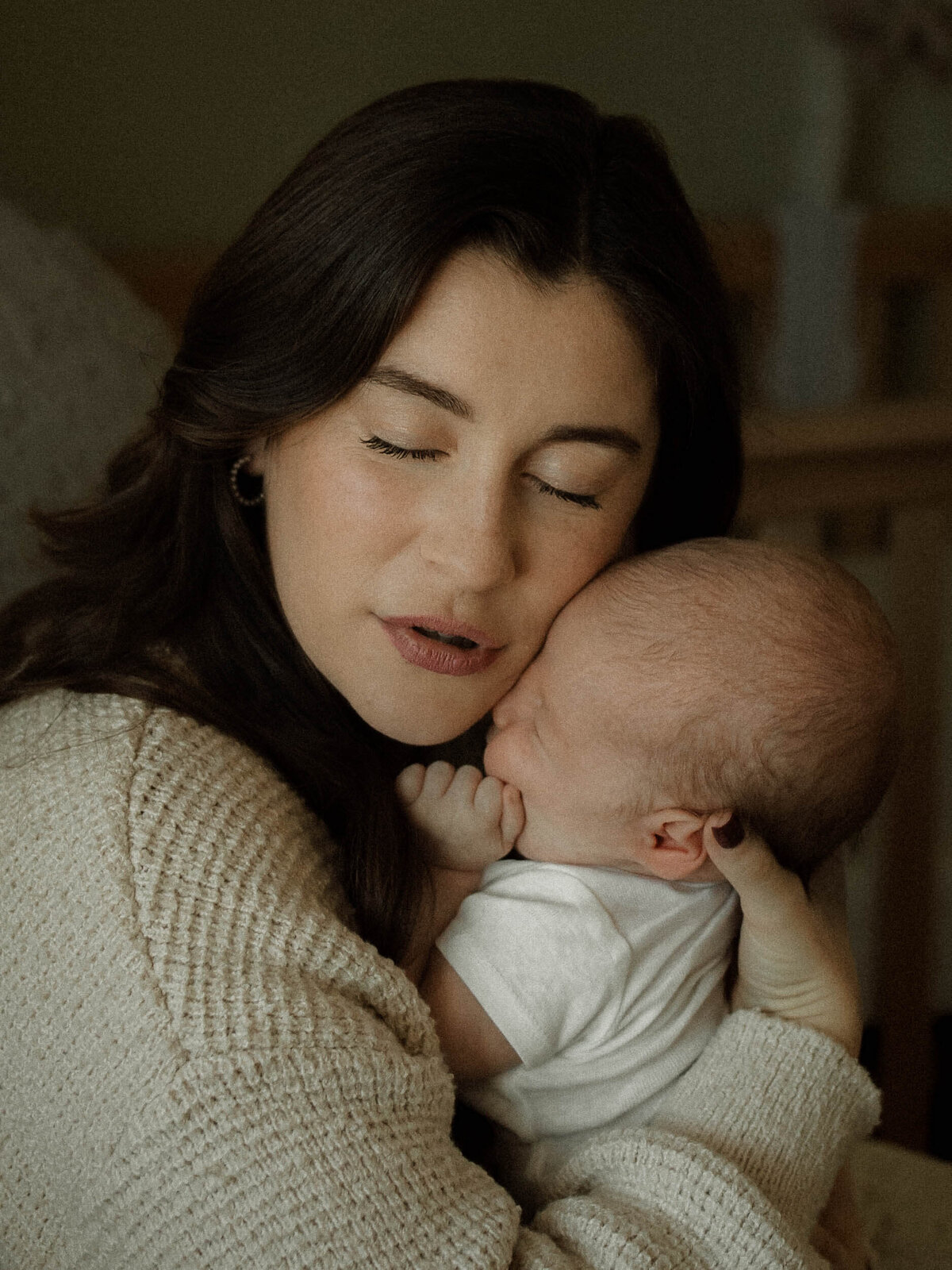 Mom holding tight her newborn son against her cheek with her eyes closed lit by window light.