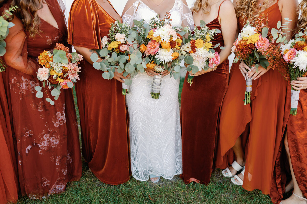 Bride and bridesmaids holding bouquets of orange, white, and greenery, bridesmaids in mismatched rust-colored dresses.