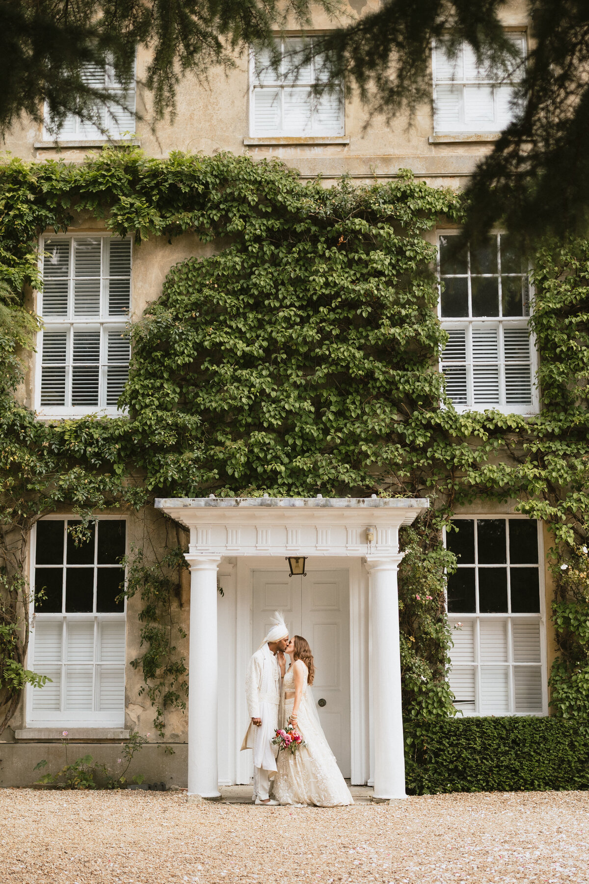 A couple stands under a white portico of a vine-covered building at Northbrook Park. The groom, in a white suit and turban, shares a tender kiss with his bride in her long white dress holding a bouquet. The charming venue features many windows with elegant shutters.