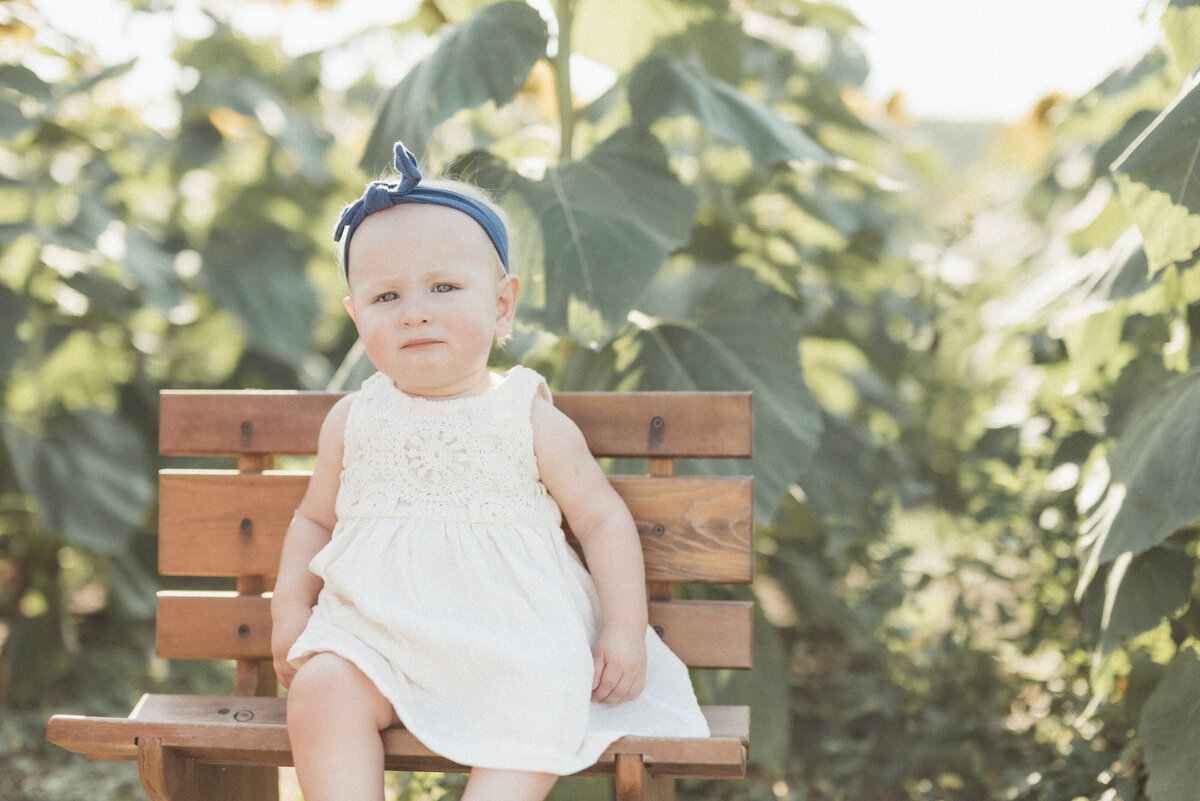 Toddler girl with a blue headband sitting on a wooden bench surrounded by green plants during a milestone photo session.