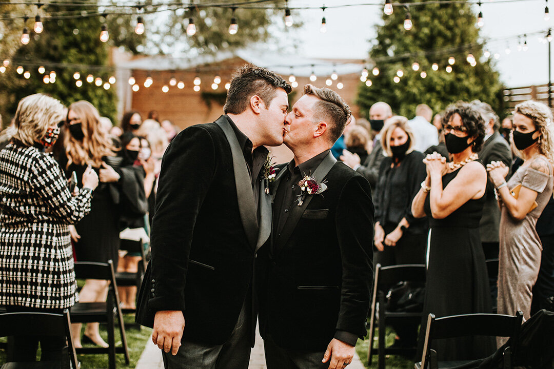 Two grooms wearing black tuxedos share a kiss at their wedding while guests look on in the background.