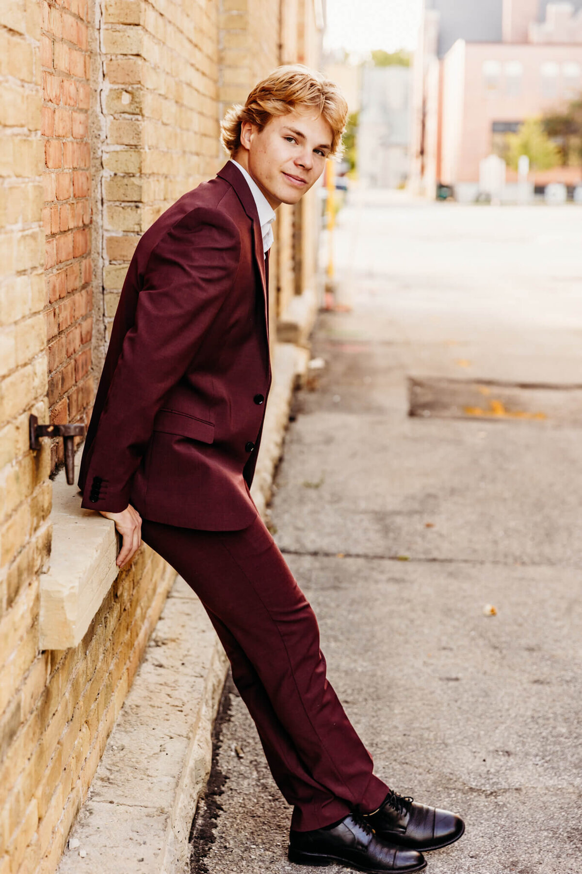 handsome teen boy in a suit sitting on a ledge in an alley while leaning forward for his senior photography session.