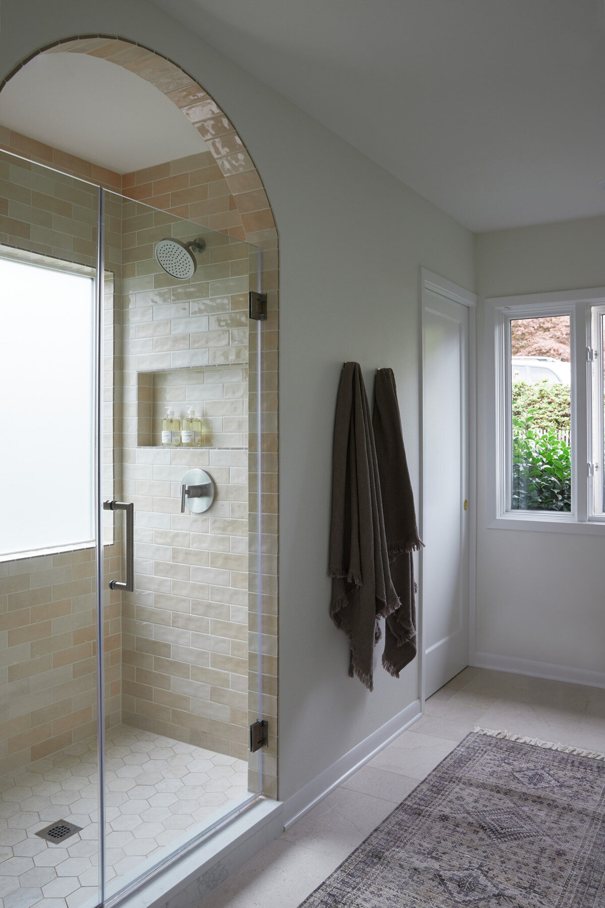 Bathroom with a shower with an arched entry and glass door. The shower has silver fixtures and a shampoo niche. The tile is brick set subway tile that’s beige and salmon in tone. To the right of the shower are two towels hooks with brown towels draped on them. A window in the right corner.