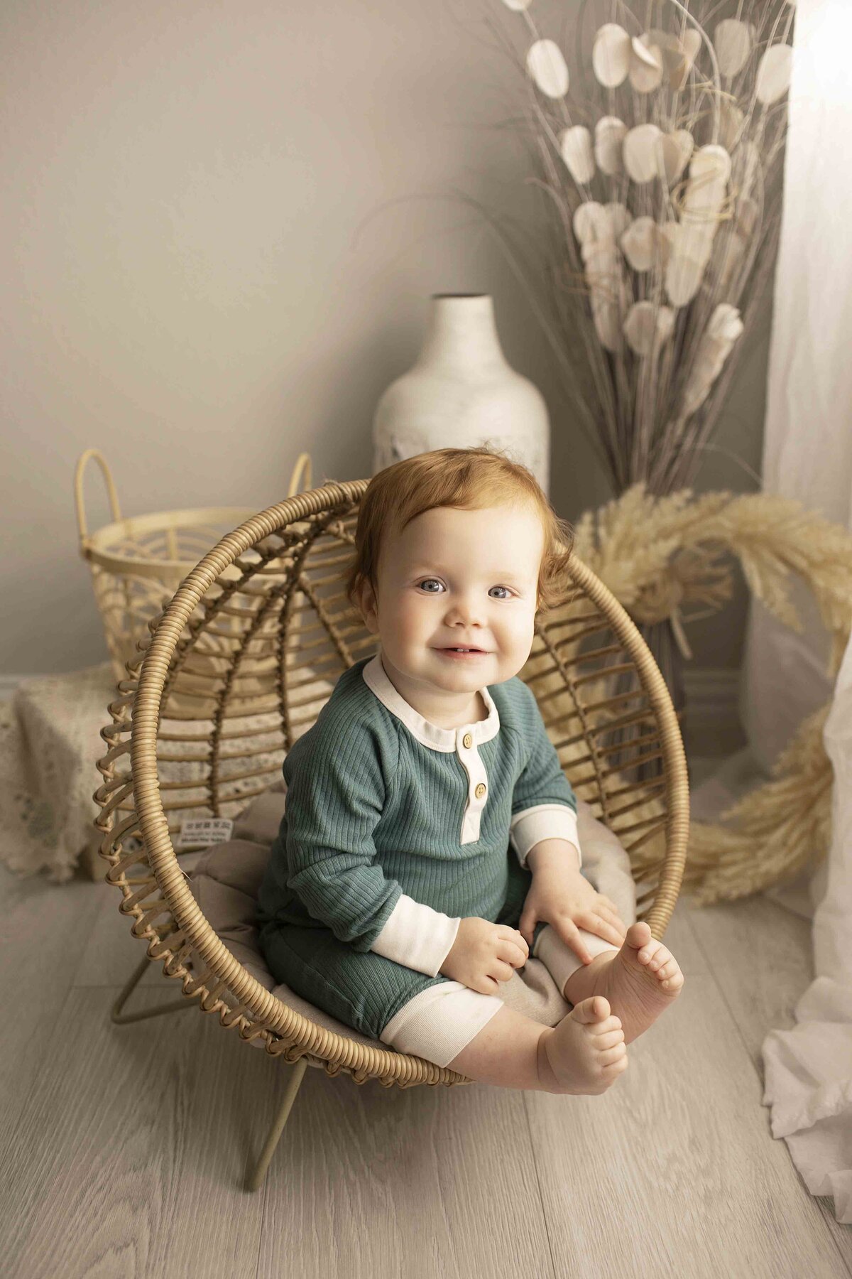 Birthday boy on his first birthday, sitting in a wicker chair, looking curiously at the camera in a soft, studio-lit space