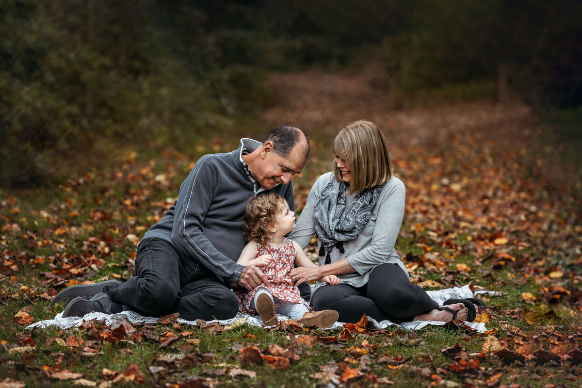 grandmother and grandfather tickling their granddaughter while sitting on a blanket surrounded by the fall leaves
