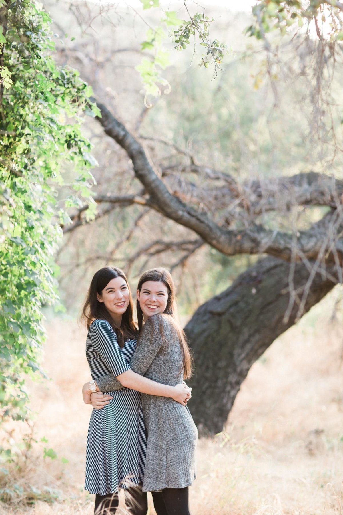 Two sisters hug it out during family photo session in a park in front of a large tree