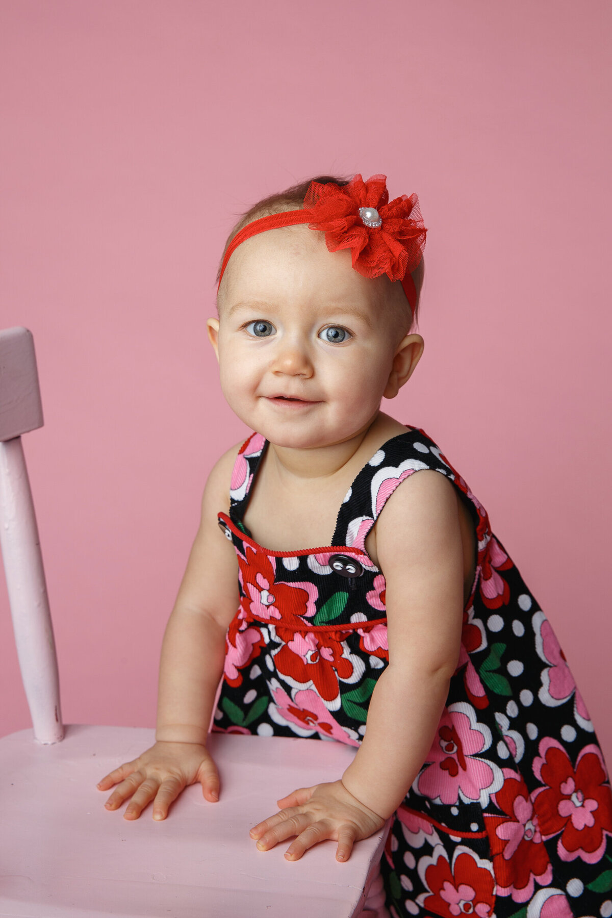 Nine month old toddler holding onto a pink chair for support  being photographed on a pink background