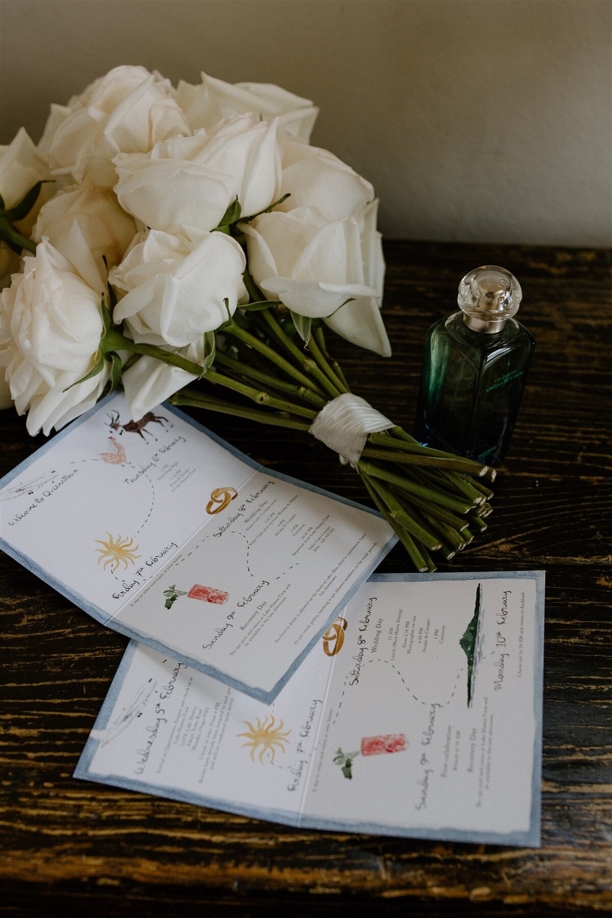 White rose bouquet beside whimsical hand-drawn wedding weekend itinerary cards and a bottle of green perfume on a wooden table.
