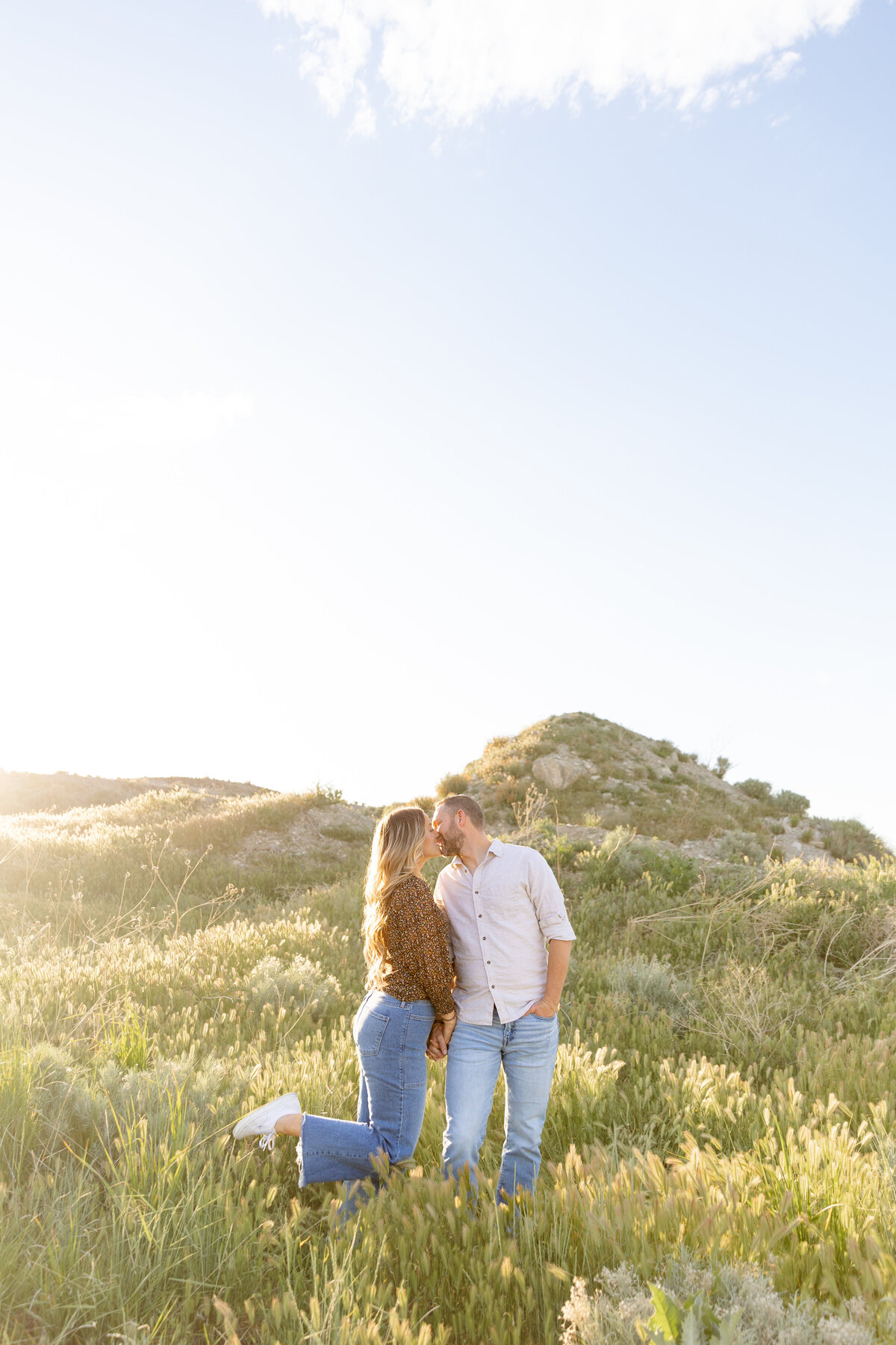 A couple shares a sweet kiss in a sunlit meadow, surrounded by rolling hills in Saratoga Springs, Utah. The woman lifts one foot off the ground as they hold hands in a playful pose.