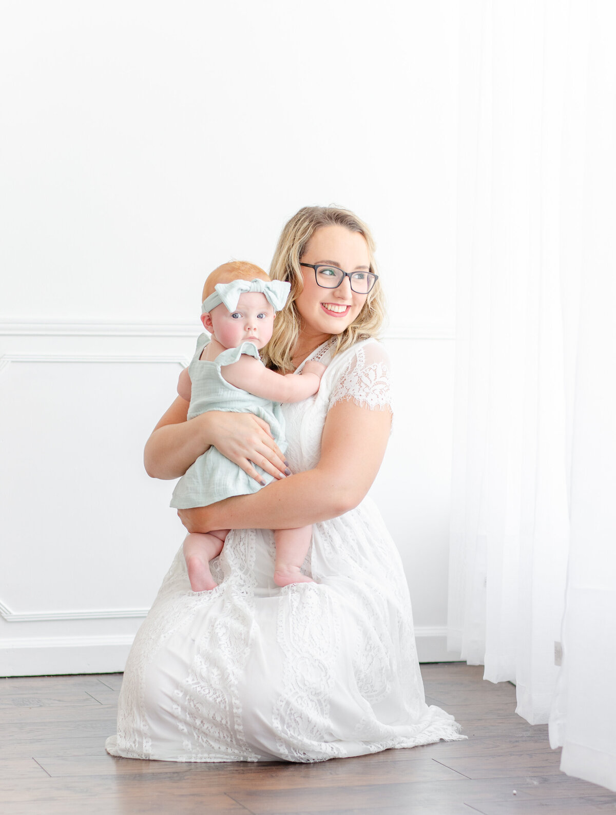 mom and baby kneel on floor next to window