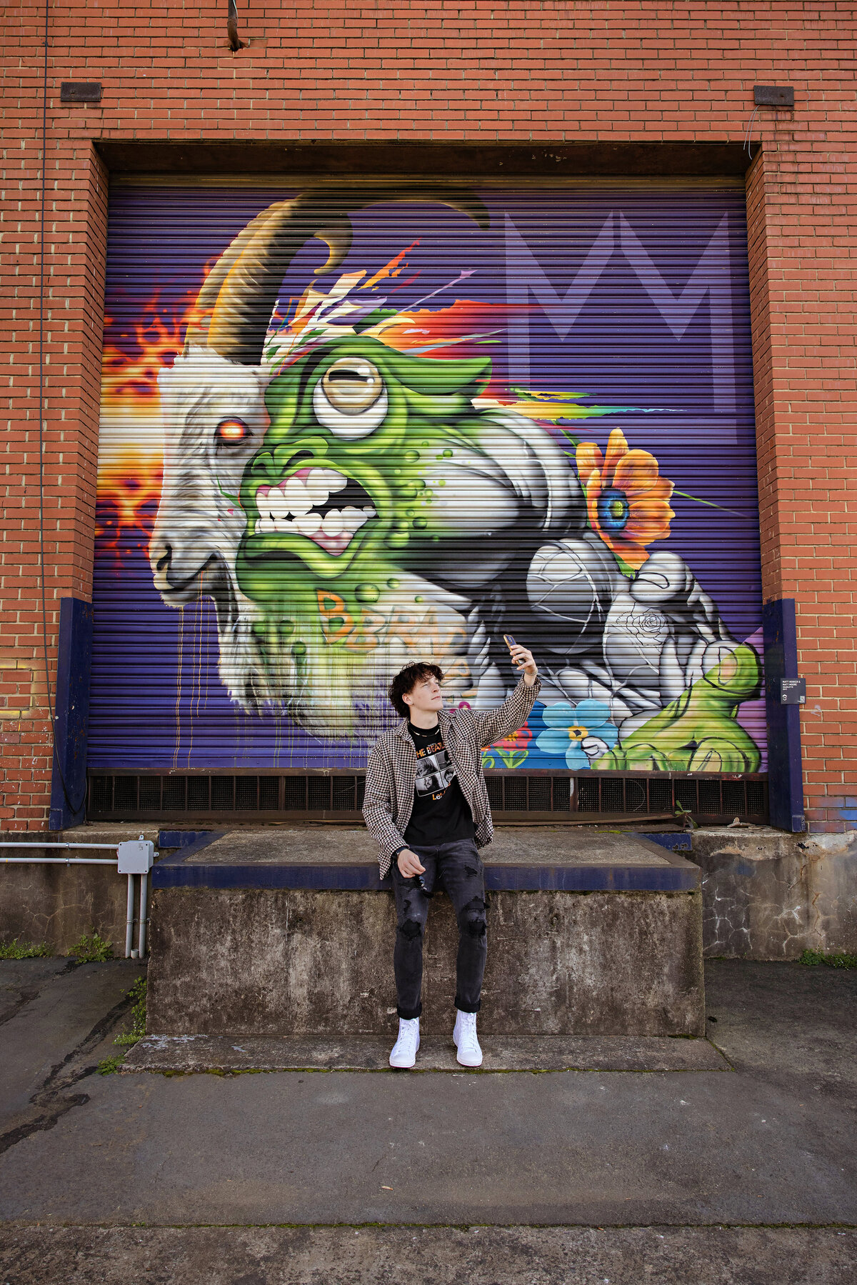 High school senior wearing a black Beatles t-shirt and checked button down at a mural-lined loading dock taking a selfie in Camp North End, Charlotte NC.
