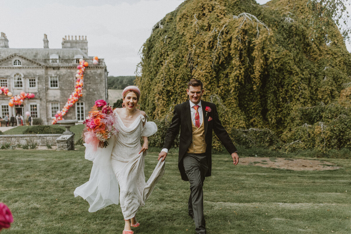 Bride with pink and orange bouquet running with groom through lawn