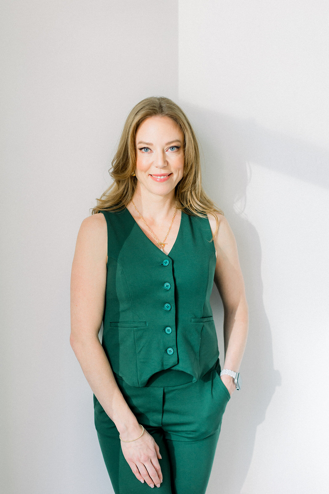 Branding photo of Calgary business woman with red hair in a greeen vest and pants leaning against a white wall