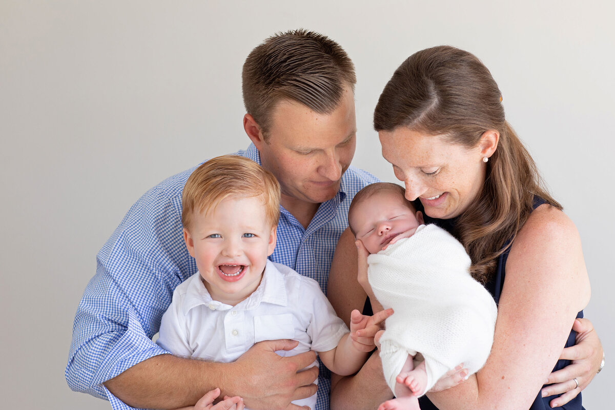 A happy family posing together, with a young child smiling beside their parents, who are lovingly holding a newborn baby wrapped in a blanket.