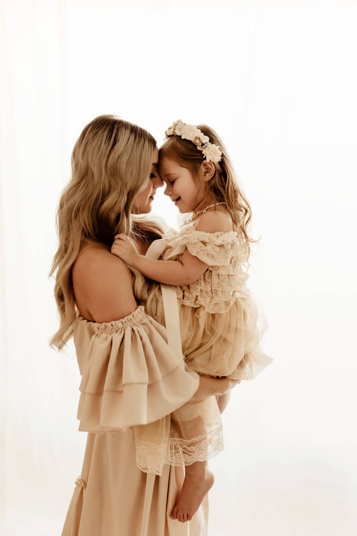 Mother and daughter touch foreheads with a white backdrop for a maternity session.