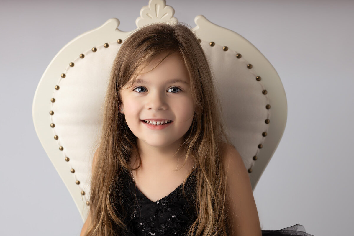 A smiling young girl with long hair in a black dress with sequins, sitting in a vintage chair.