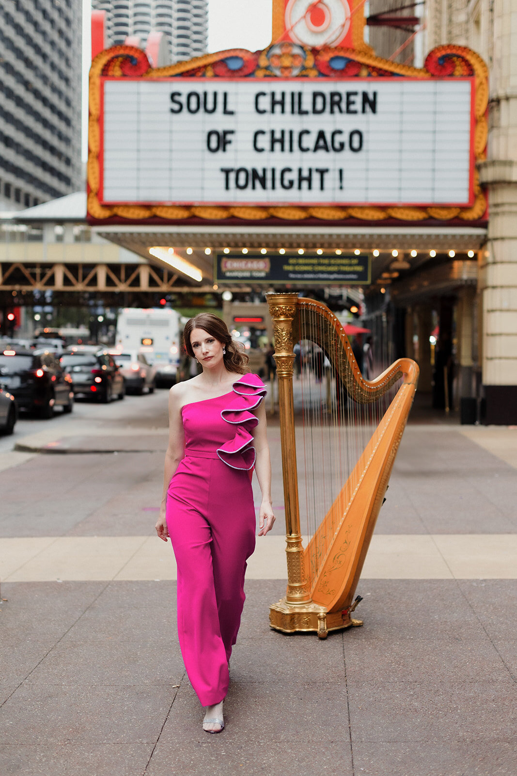 woman in hot pink jumpsuit is walking toward camera with her harp and Chicago Theater in the background.