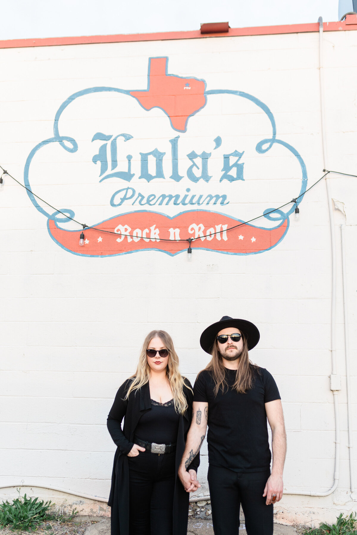 A couple posing seriously in front of a music venue during their engagement session in Fort Worth, Texas. The woman on the right is wearing a black top, black pants, a long black jacket, and sunglasses. The man on the right is wearing a black shirt, black jeans, a black rimmed hat, and sunglasses.