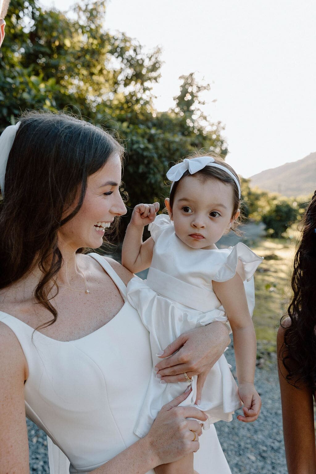 Bride holding and smiling at little flower girl outside with trees behind them