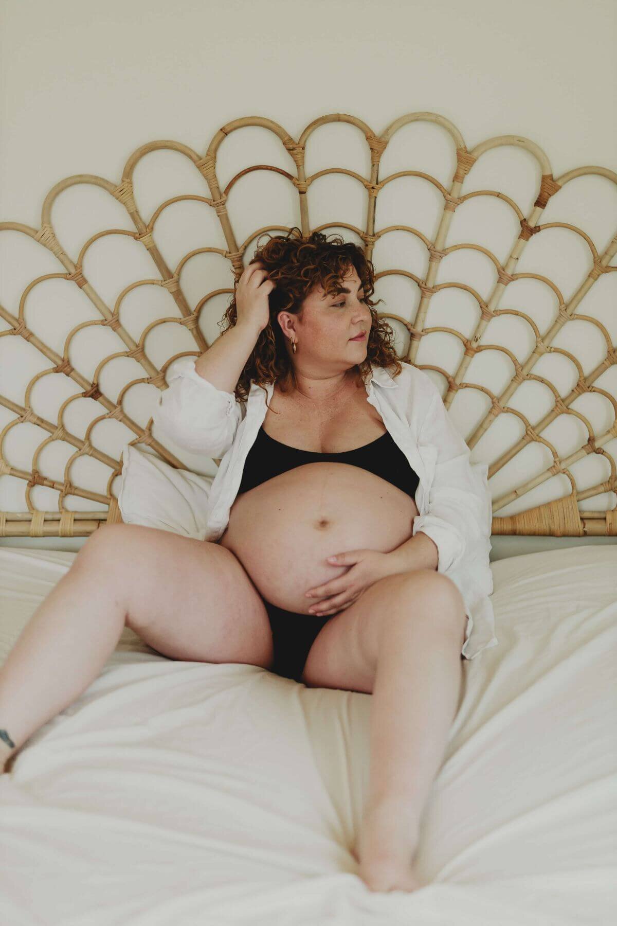 Gravidbild på en gravid kvinna sittandes på en säng med vita lakan under en gravidfotografering i Stockholm