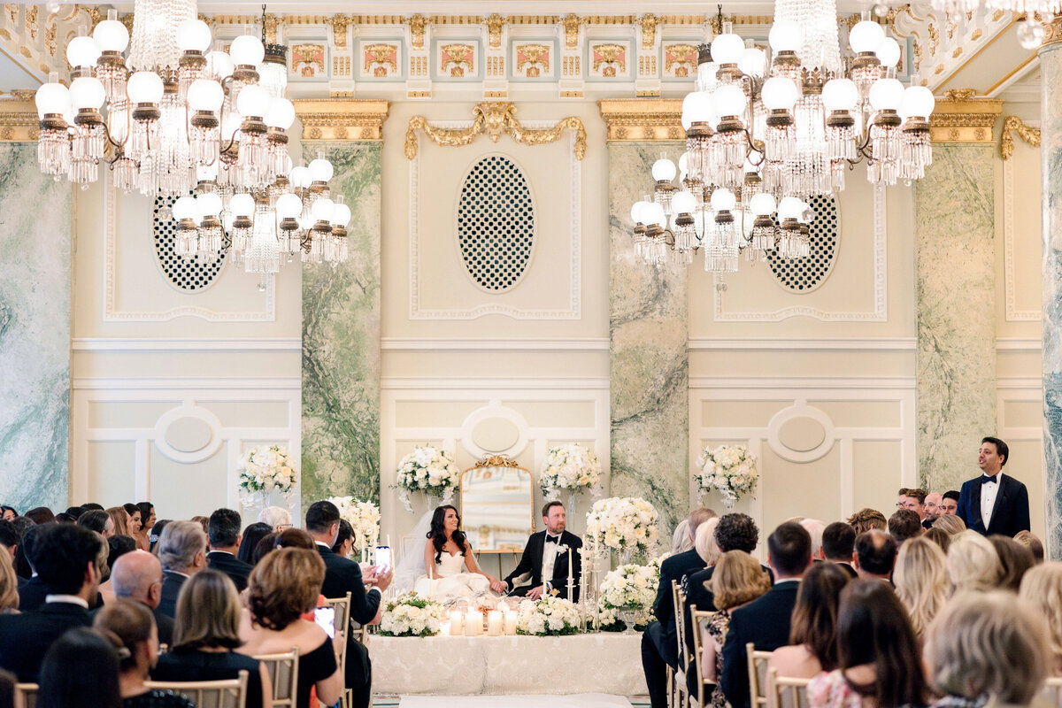 A bride and groom sit at a lavishly decorated table during their wedding ceremony in an elegant, ornate hall. The room is filled with guests and features large chandeliers, marble walls, and intricate details, creating a luxurious atmosphere.