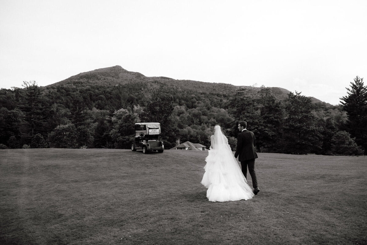 The bride and groom walk toward the golf cart after the wedding ceremony at The Ausable Club, NY, with mountains in the background.