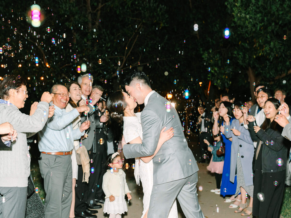 Bride and groom kiss under floating bubbles, surrounded by joyful guests at night. Guests smile, capturing a celebratory and romantic atmosphere.
