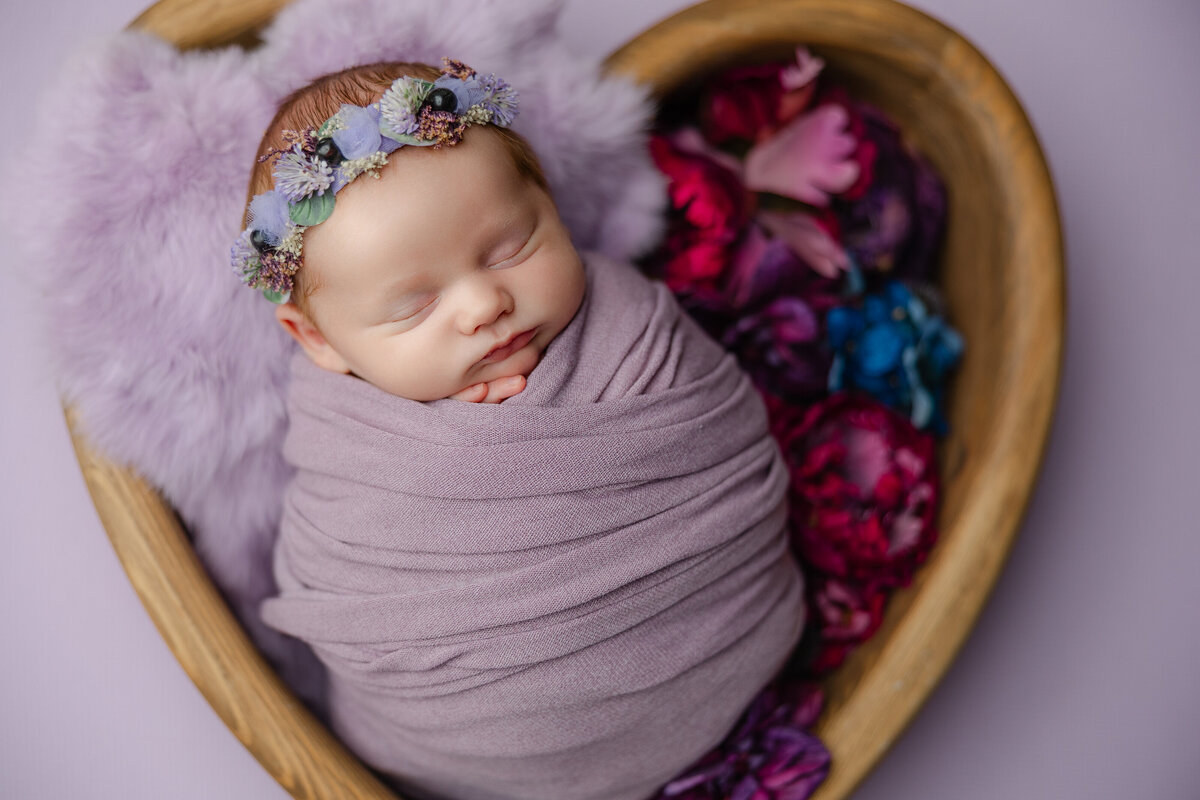 Baby girl swaddled in mauve with a pastel flower headband, nestled beside pink and blue florals on a soft fur layer. Photographed in-studio by Inland Empire newborn photographer Kaitlyn Dawn.
