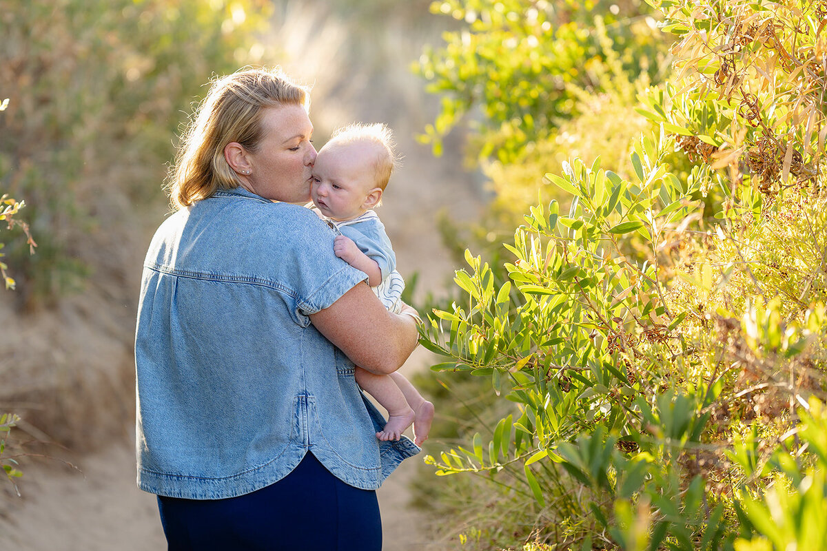 Natural light motherhood portrait in Geelong showing a mother kissing her baby while walking through a sunlit bushland trail.