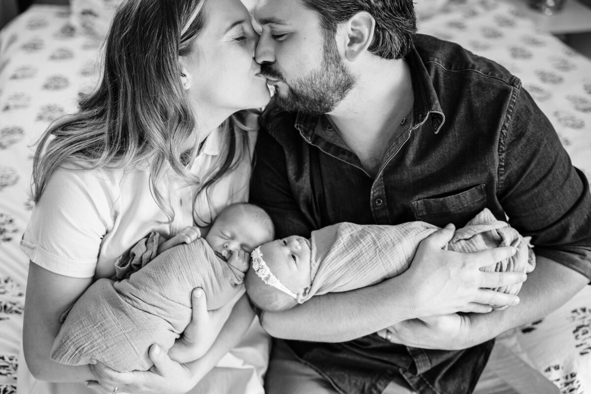 A black and white photo of a couple tenderly kissing while holding their newborn twins, one in each arm.