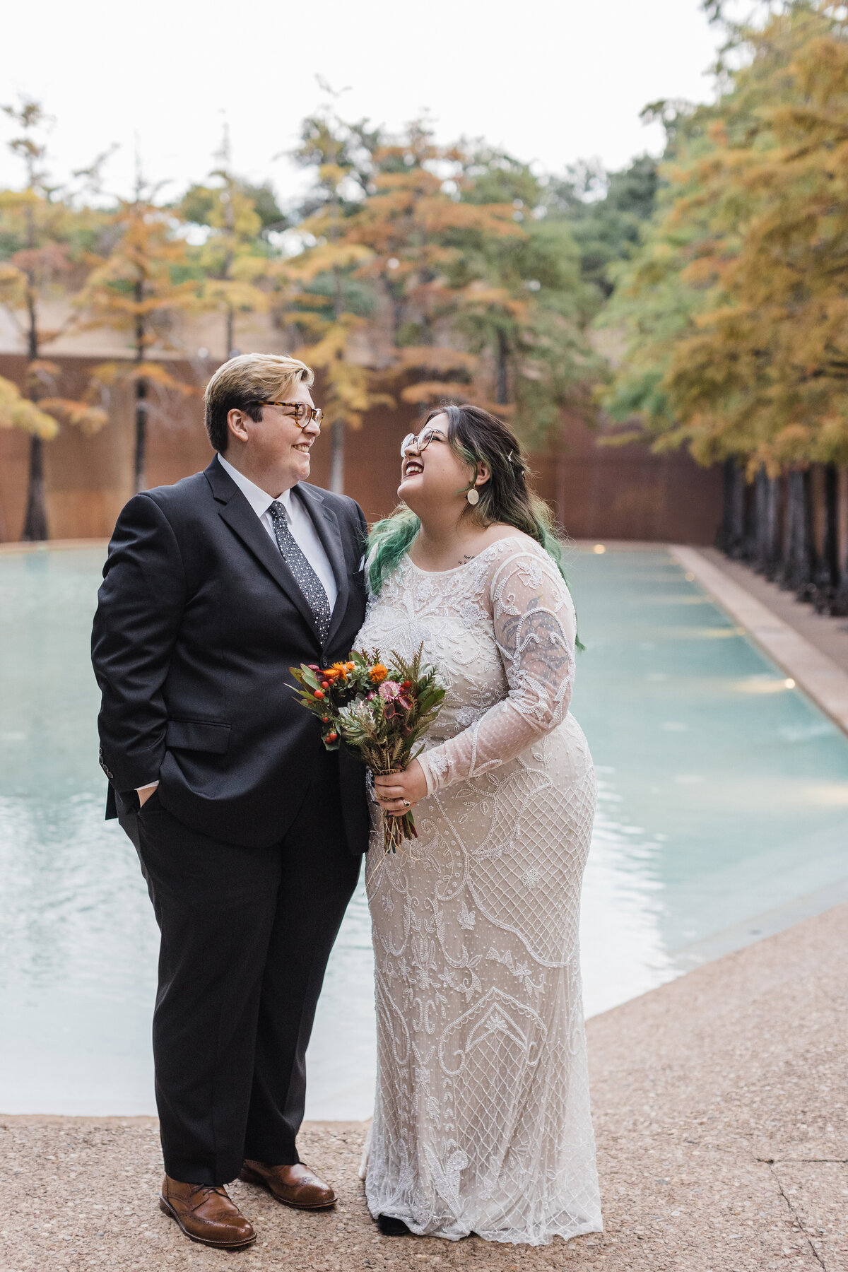 A bride and a marrier posing together after their elopement ceremony at the Fort Worth Water Gardens in Fort Worth, Texas. The bride is on the right and is wearing an intricate, long sleeve, white dress and is holding a bouquet. The marrier is on the left and is wearing a dark suit with a tie.