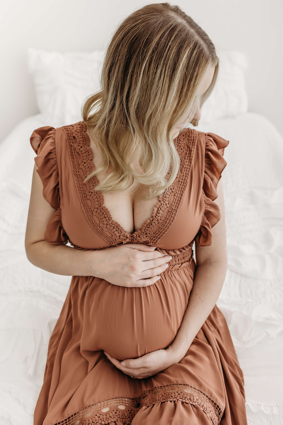 Mom in rust colored dress sitting on bed holding pregnant belly