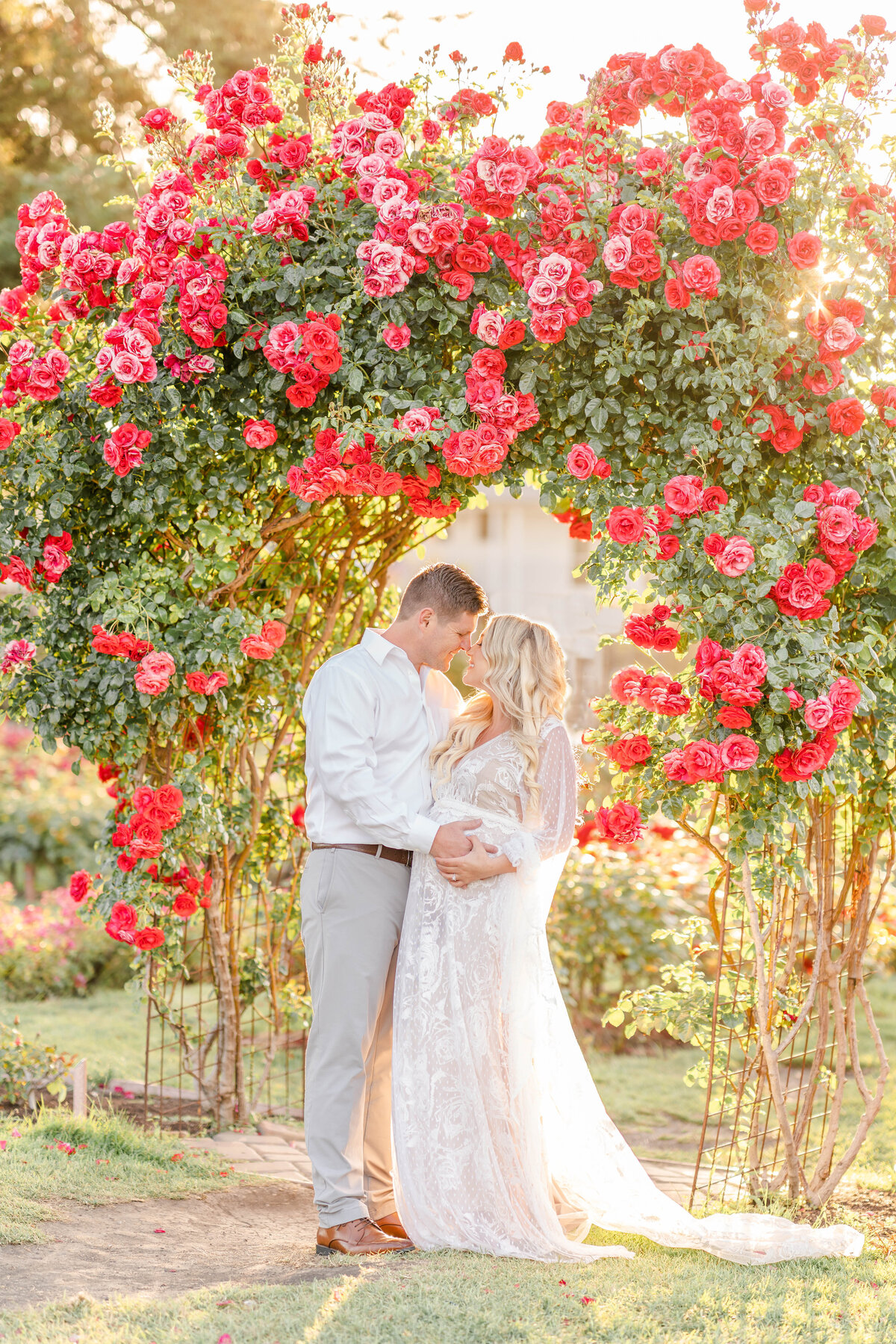An expecting couple stands in a rose garden archway looking into one another's eyes while both caressing her beautiful baby bump photographed by Bay area photographer, Light Livin Photography.