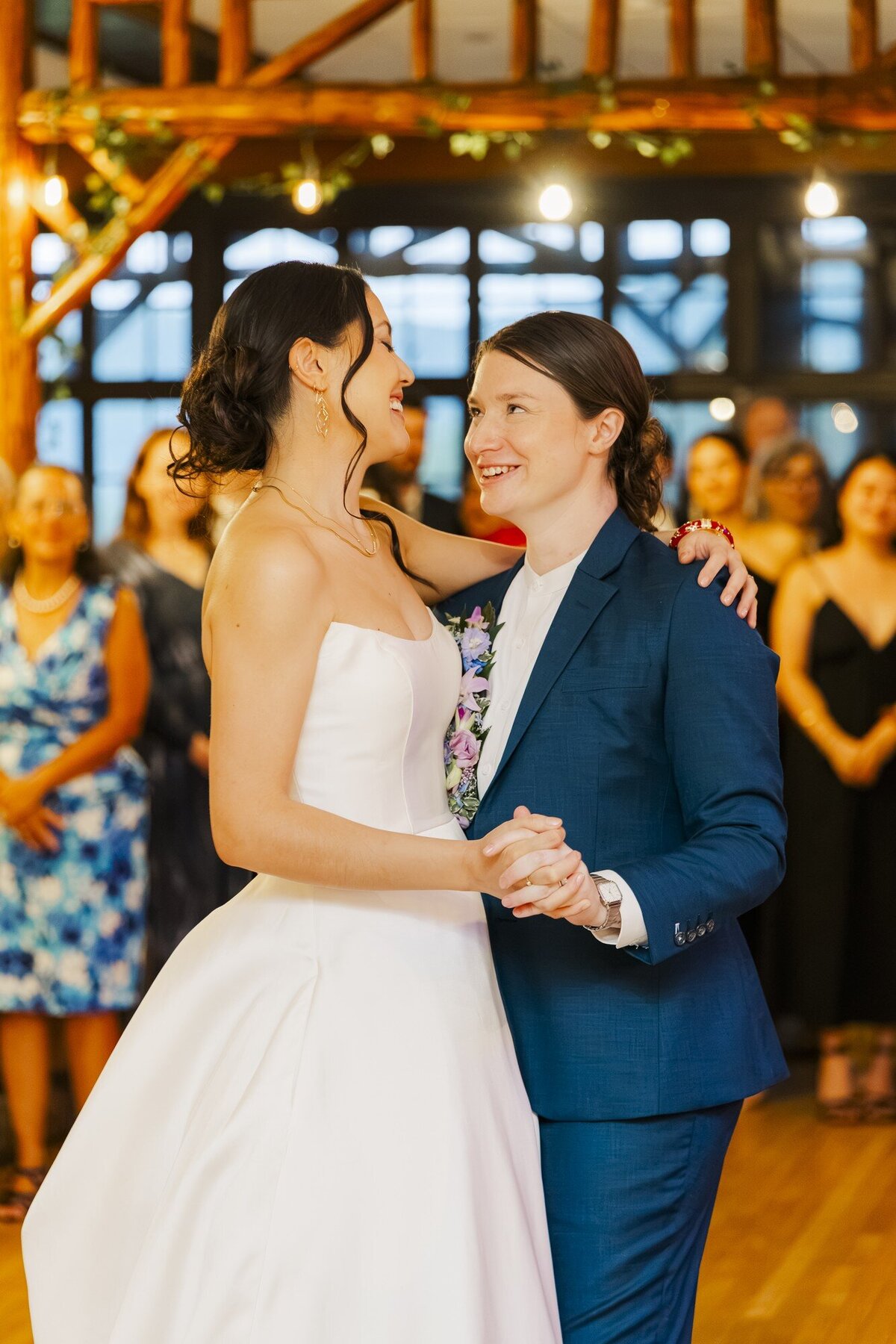 Two brides slow dancing in a barn reception, one in a gown and one in a blue suit