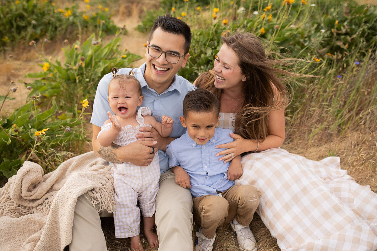 Parents and young children laugh together while sitting on a blanket in a field with yellow and purple flowers. 