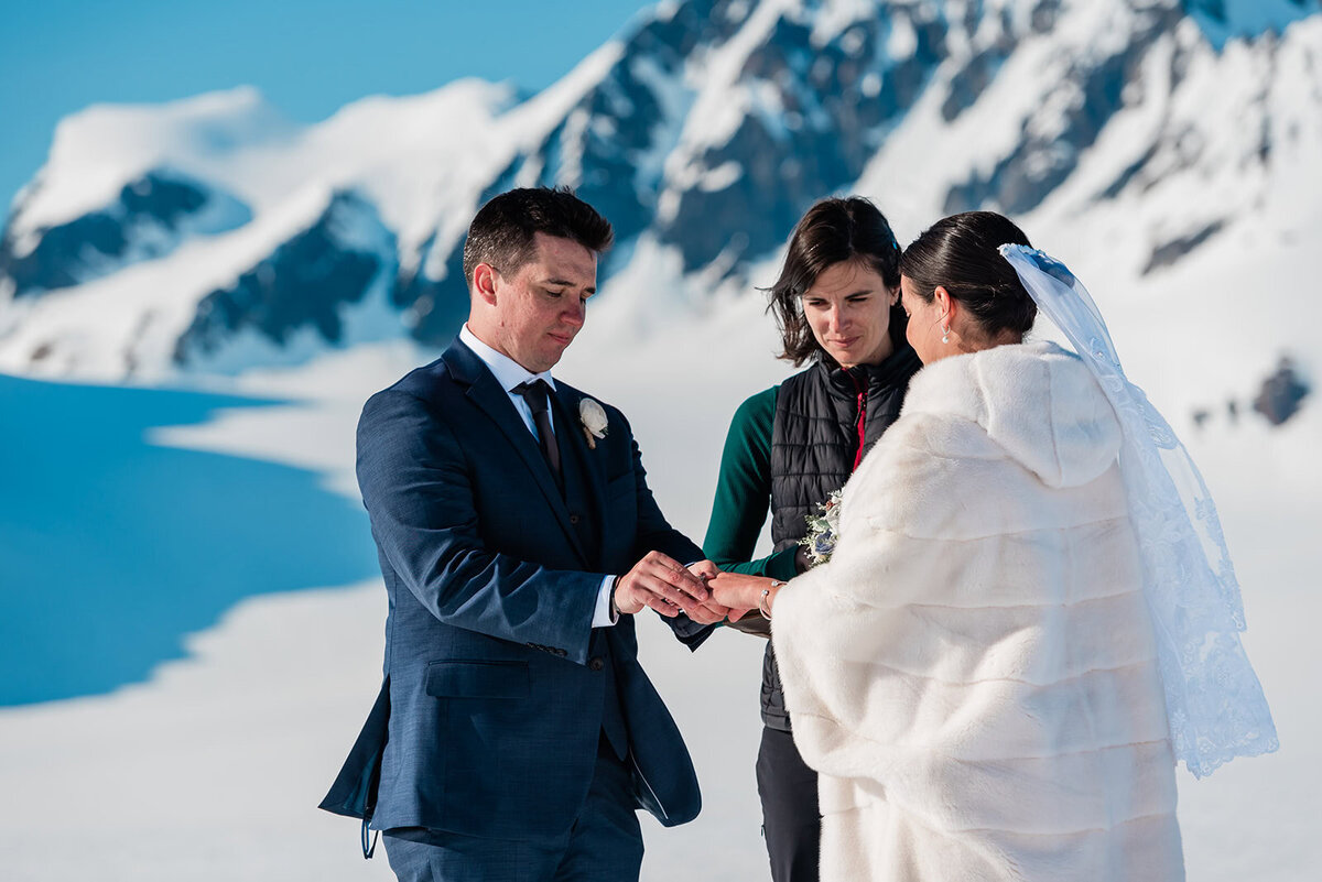 A bride and groom in their wedding finest exchange rings with their officiant guiding them during elopement vows on the glacier during their two day adventure luxe Alaska elopement.   The sky is bright blue and the snow gleams white against the dark mountains rising up from the glacier.