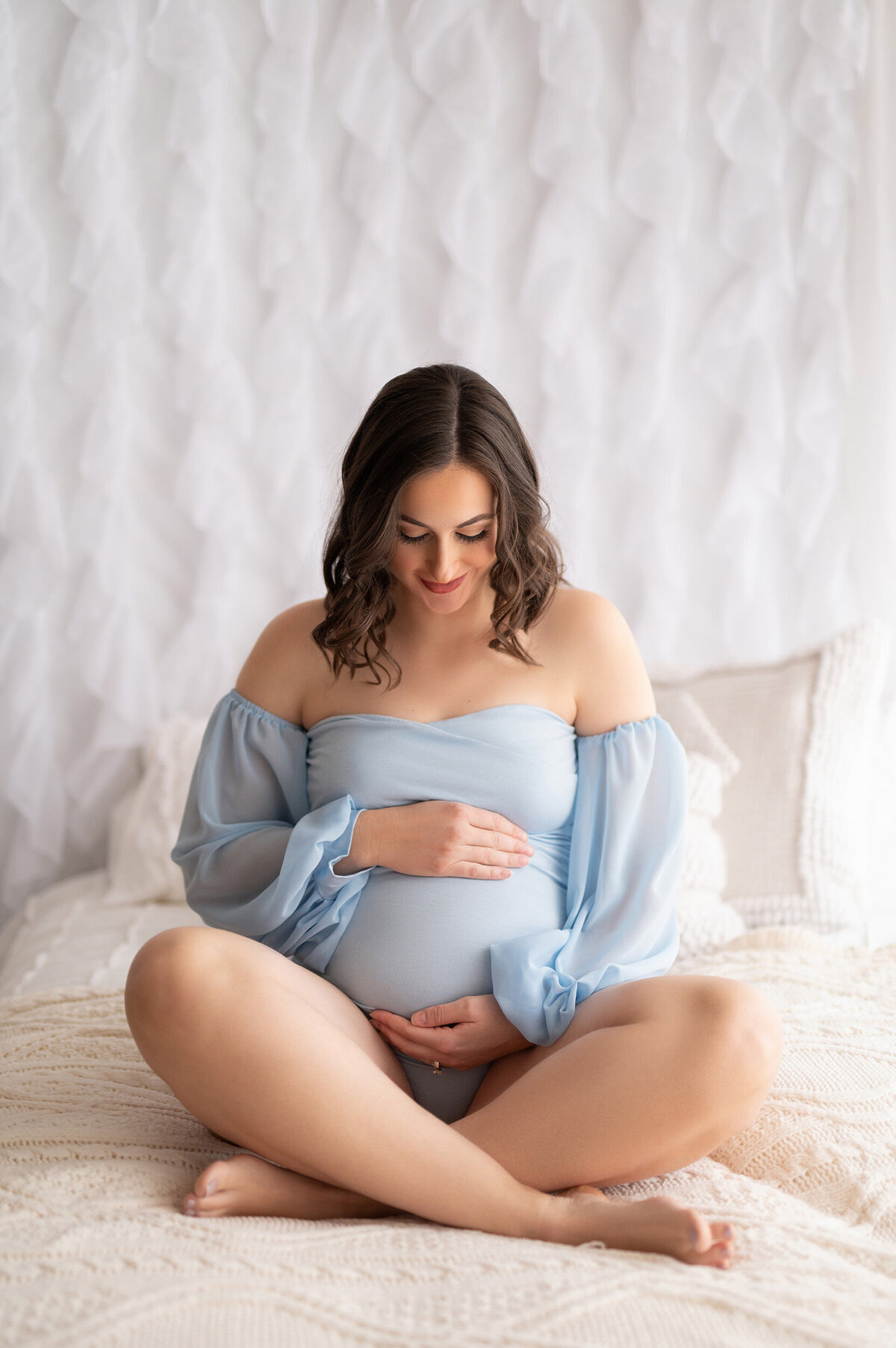 Pregnant woman sitting cross-legged on a bed facing the camera but looking down.  She is cradling her belly while wearing a sheer, peasant sleeved, maternity bodysuit.