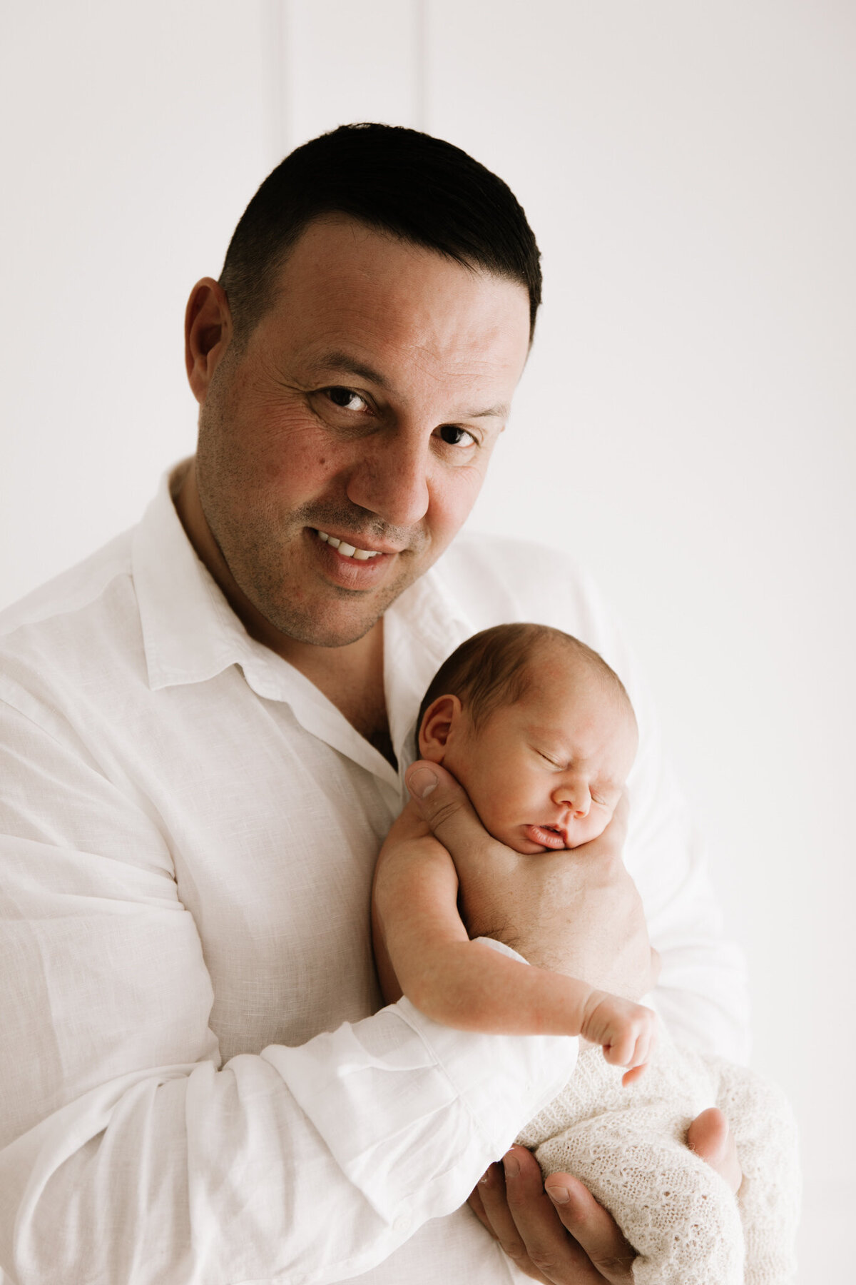 Father in a white shirt smiling while holding his newborn baby gently in his hands during a softly lit session in a Southwest Sydney photography studio.