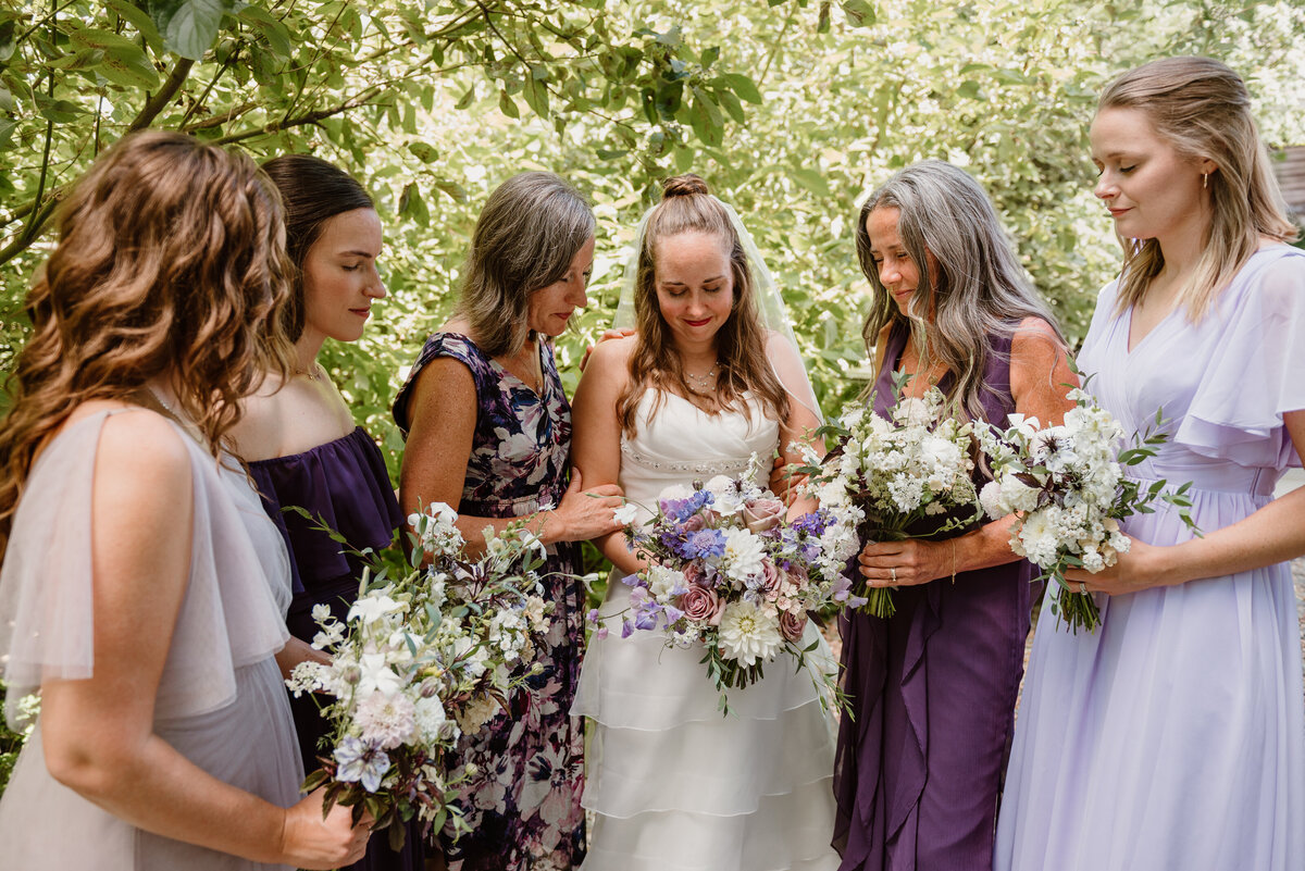 Bridal party in Bozeman, Montana surrounded by summer greenery, holding lush hand-tied bouquets in soft lavender, cream, and purple tones. Elegant garden roses, lisanthus, designed by Indigo & Violet Co., for a romantic outdoor wedding. 