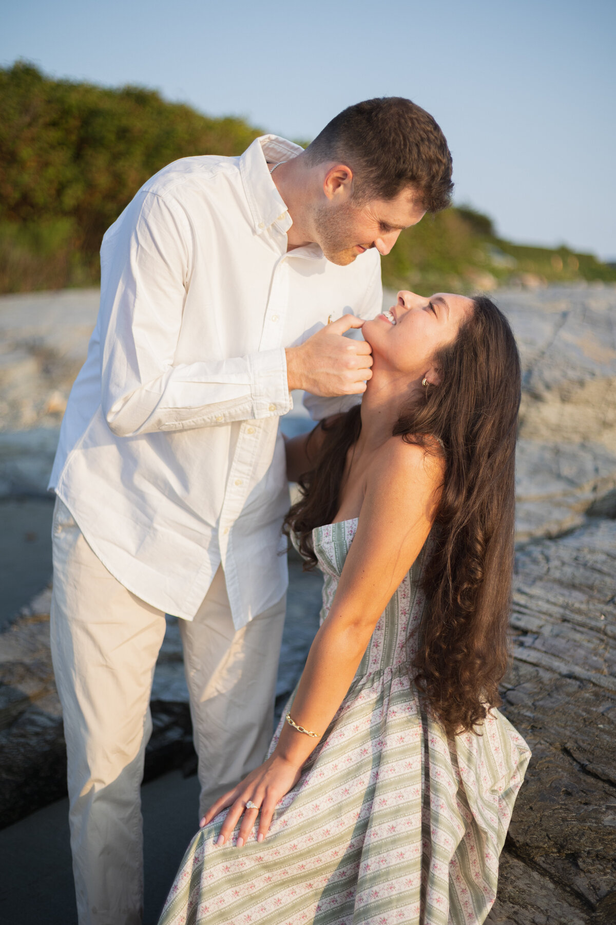 Collins Beach Newport RI | Kelsey Sheehan Photography Timeless Rhode Island Weddings | A couple shares a tender moment at the beach. The man, in a white shirt, gently tilts the laughing woman's chin upward. The sun casts a warm glow.