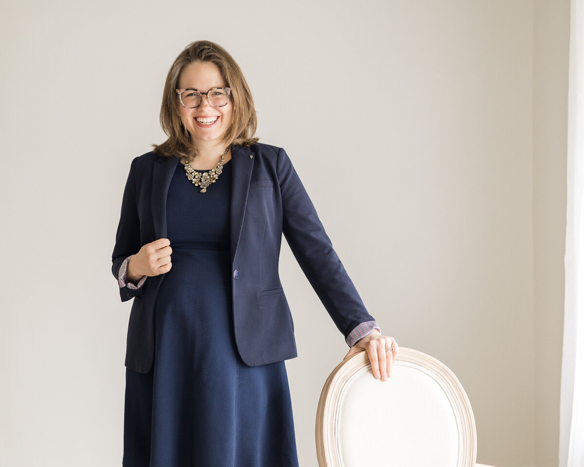 Brunette blonde pregnant female standing with her hand on a chair, wearing glasses, a dark blue dress with a long sleeve dark blue jacket, smiling at the camera