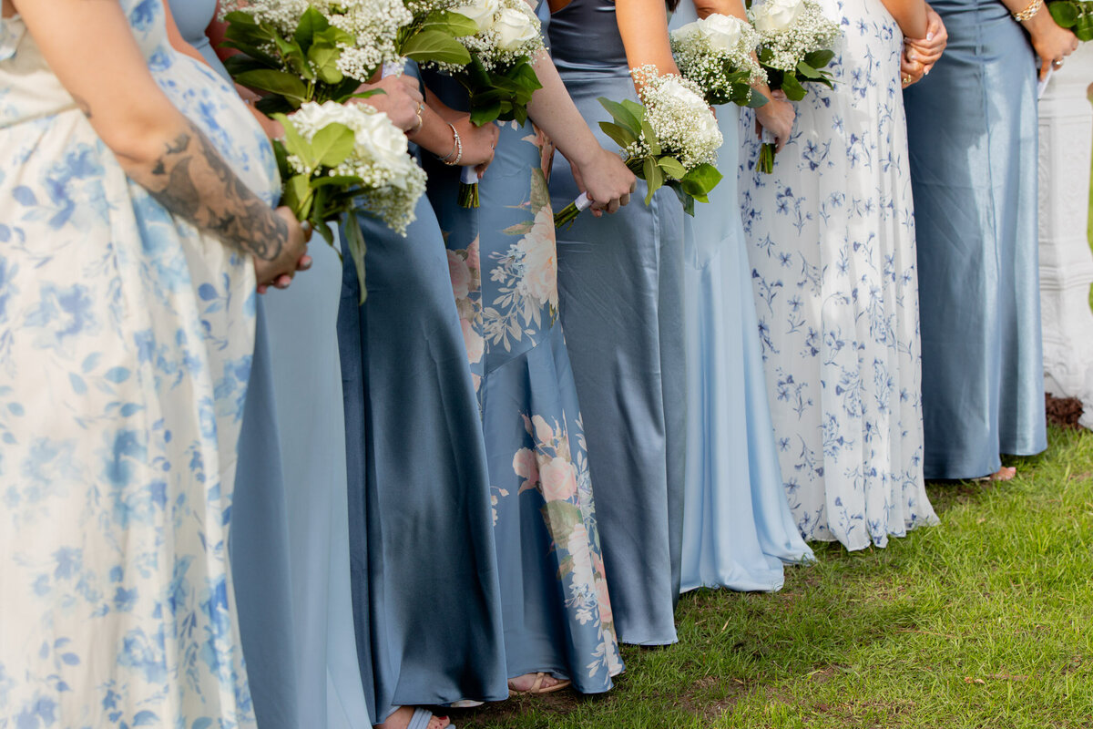 Cropped photo of the bridesmaids during the wedding ceremony. They are all wearing different dresses, different shades of blue and holding a small bouquet of white flowers.