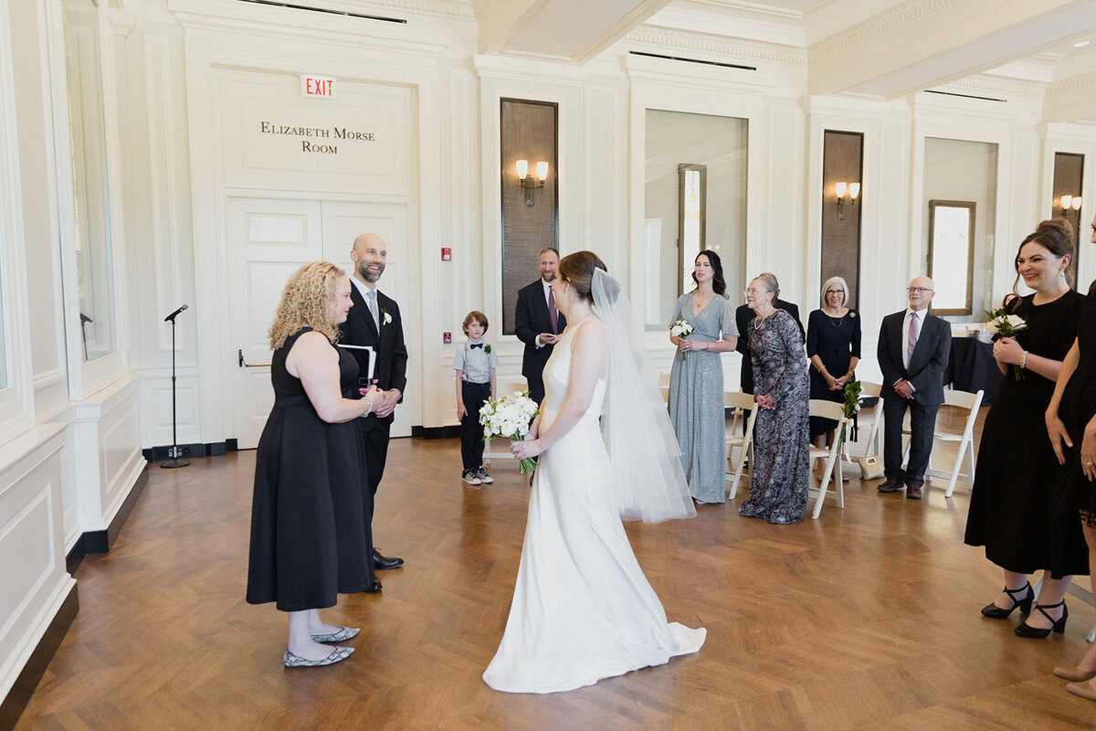 Wedding couple meets the officiant at the front of the ceremony to be married. Small number of guests are behind them.