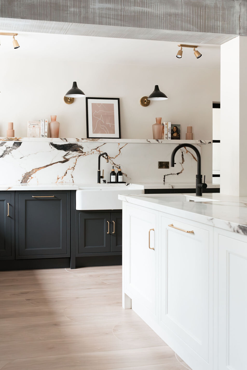 A modern kitchen with dark lower cabinets, a white marble backsplash with black and gold veining, a farmhouse sink, and minimalist open shelving holding books and decor.  A center island has matching marble counters, paired with white lower cabinets.