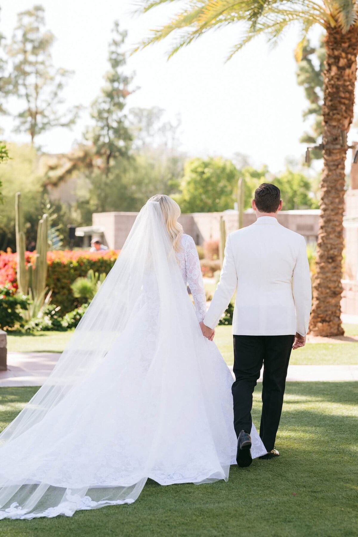 Arizona Biltmore wedding photography capturing a bride in a lace gown and long veil walking hand in hand with her groom in a white jacket across the resort gardens.