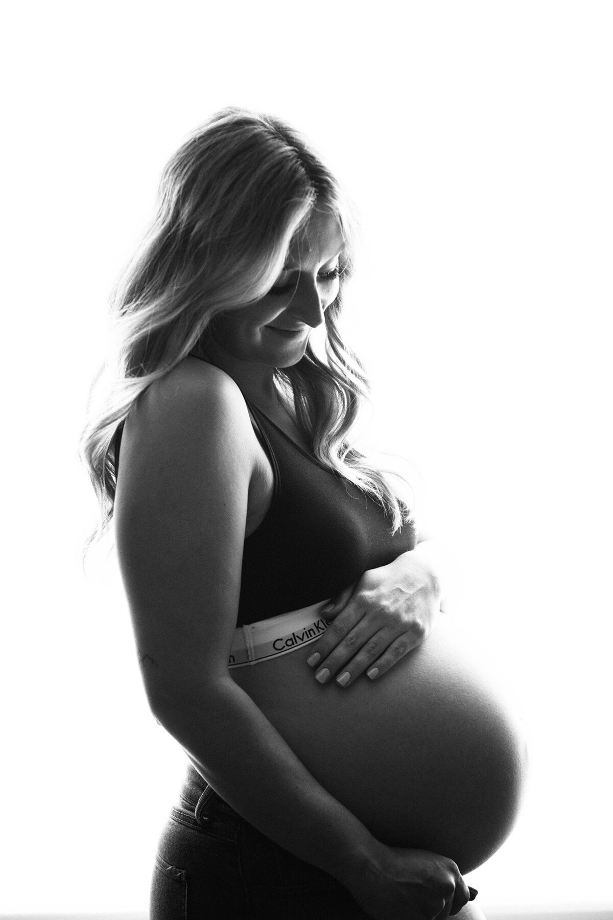 A black and white image of a mom to be smiling down to her bump while standing in a studio in a sports bra and jeans