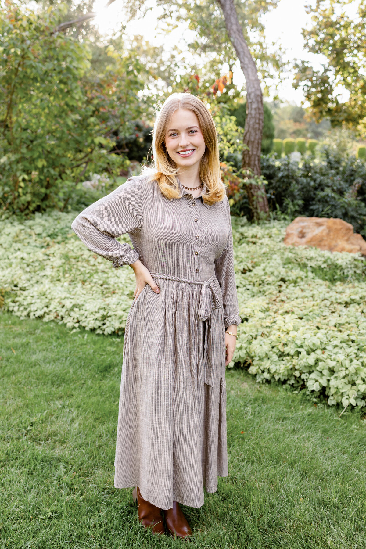 A young woman in a taupe dress poses gracefully in a well-manicured garden with vibrant greenery behind her.